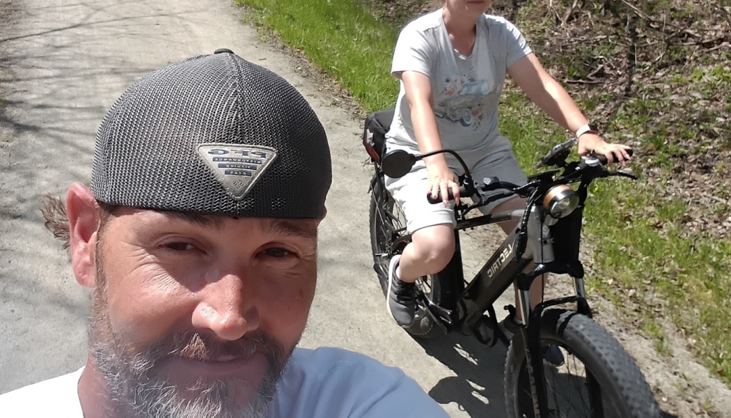 A bearded man taking a selfie while a woman rides a black electric bike on a sunny dirt path.