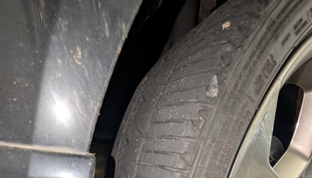 Close-up of a heavily worn car tire with a tear and damaged tread beneath a dark vehicle.