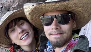 A smiling young couple outdoors wearing straw hats, with the man in sunglasses and the woman smiling warmly.