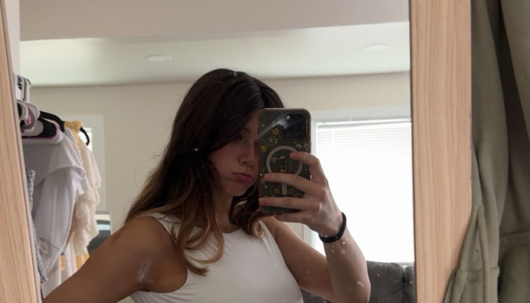 Young woman with long brown hair taking a mirror selfie with her smartphone while wearing a white top.