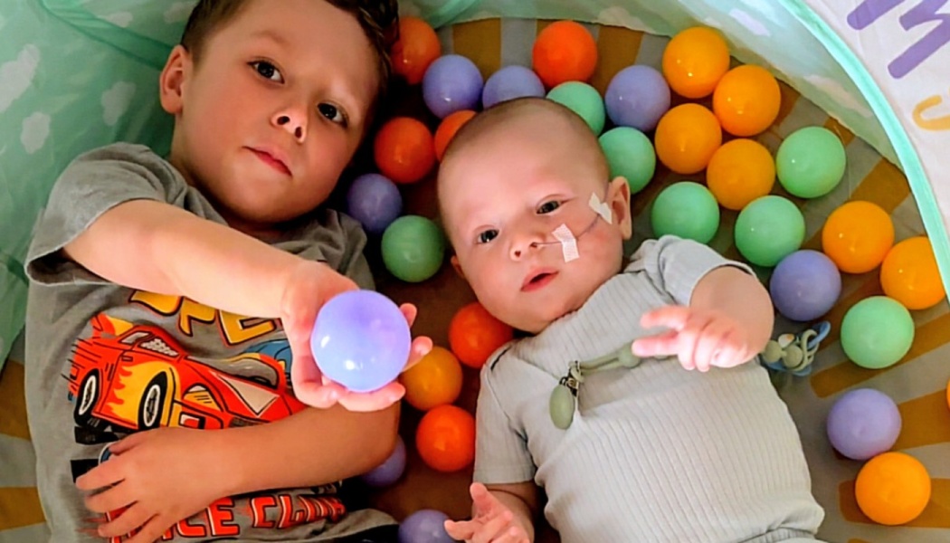 A young boy and a baby with a nasal tube lie side-by-side in a colorful ball pit.