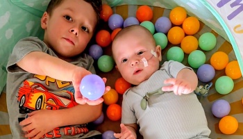 A young boy and a baby with a nasal tube lie side-by-side in a colorful ball pit.