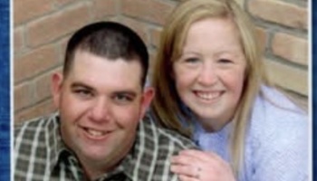 A smiling man in a plaid shirt and a blonde woman pose closely together in front of a brick wall.