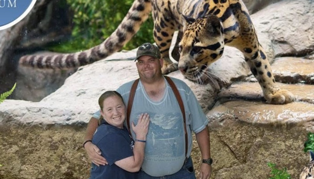 A smiling couple embraces with a large, spotted wildcat perched on rocks directly behind them.