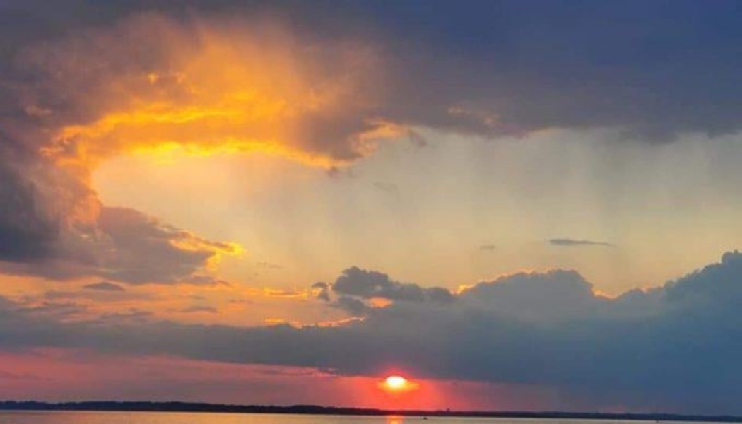 Glowing red sun setting over water beneath dramatic storm clouds illuminated with bright yellow and orange light.