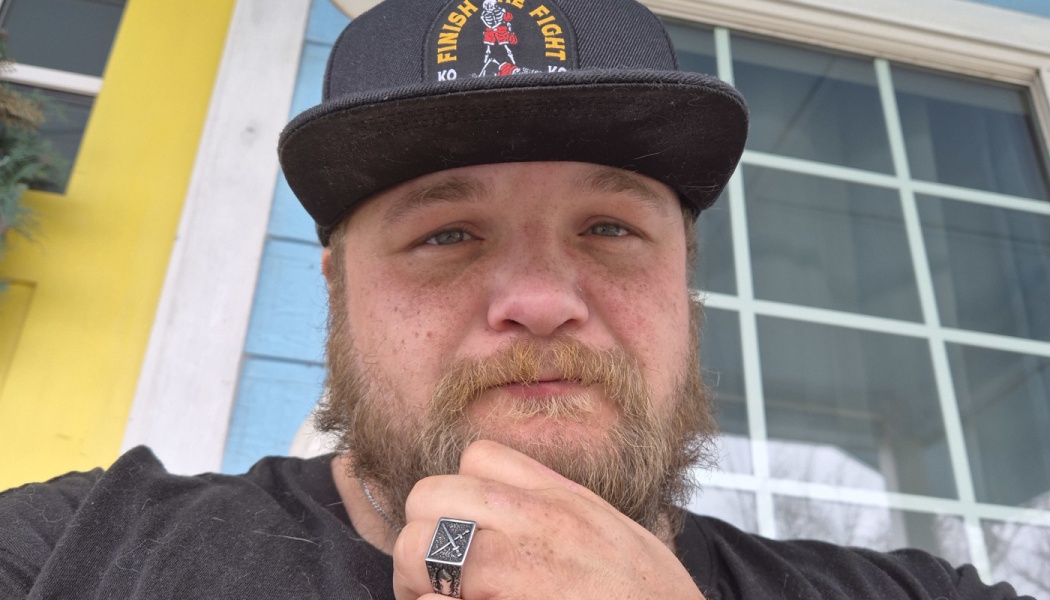 Bearded man resting his hand on his chin, wearing a black "Finish the Fight" cap and a silver ring.