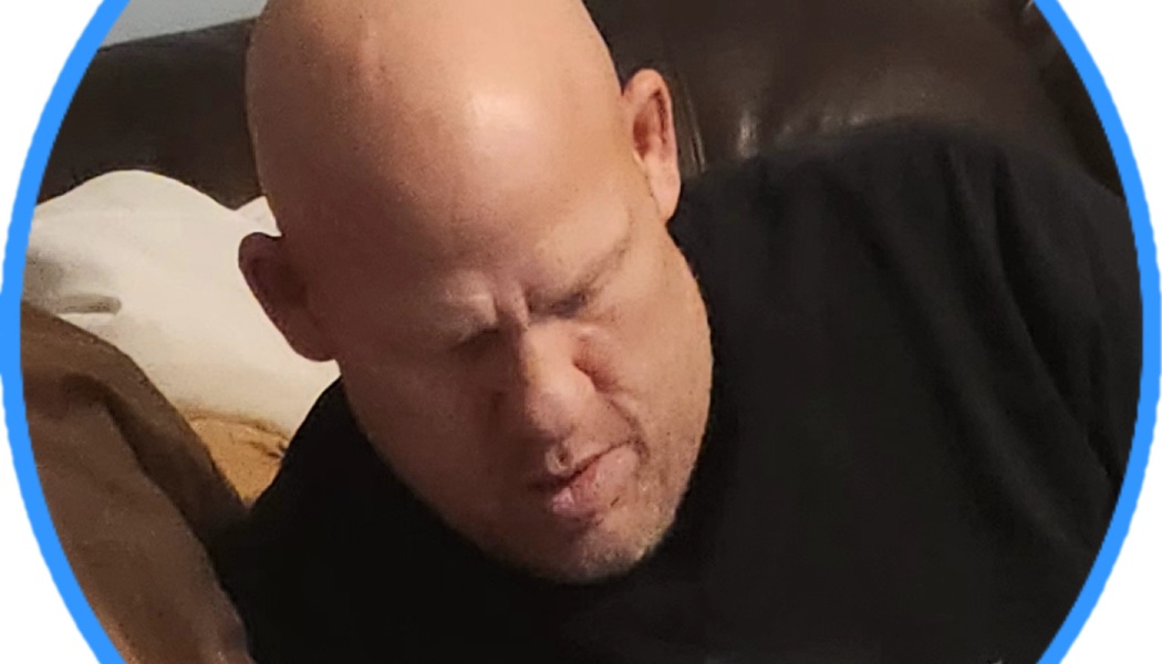 A bald man in a black shirt sitting on a couch with his eyes closed, framed in a blue circle.