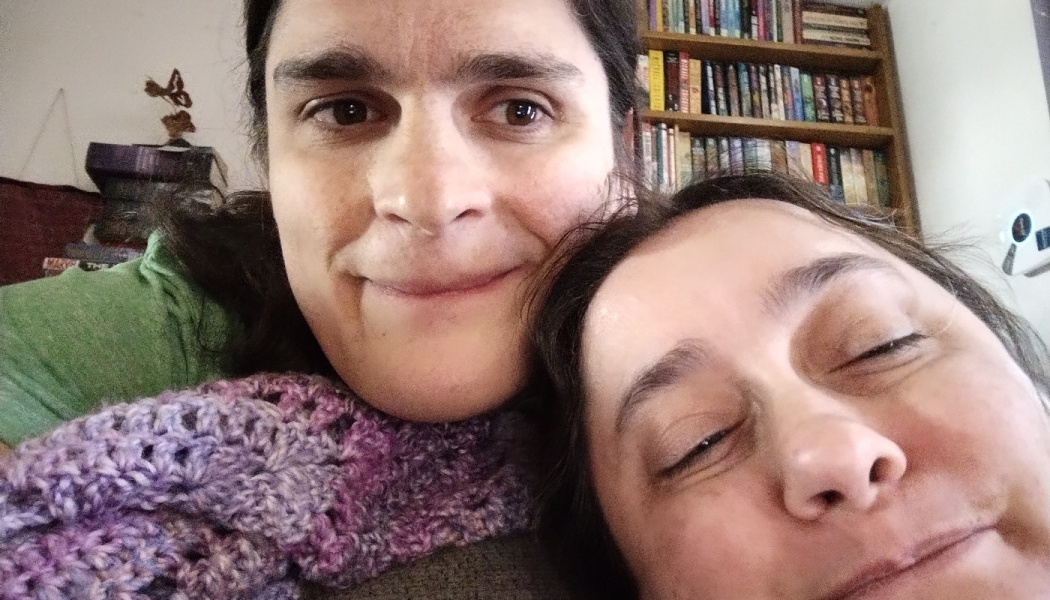 Close-up selfie of two people leaning close together indoors, with a bookshelf filled with books behind them.