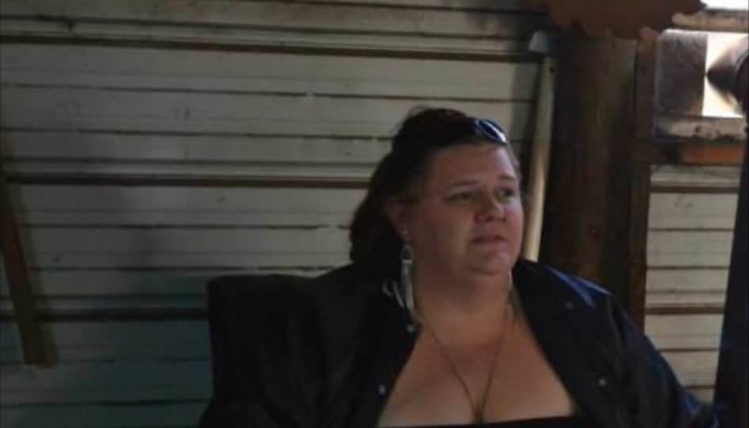 A woman with dark hair and sunglasses on her head sitting in front of a rustic slatted wall.