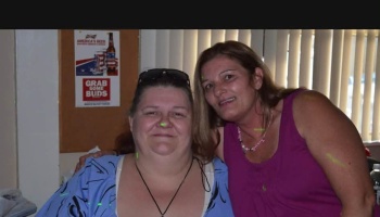 Two women posing closely together and smiling, one in a blue patterned shirt and the other in a purple tank top.