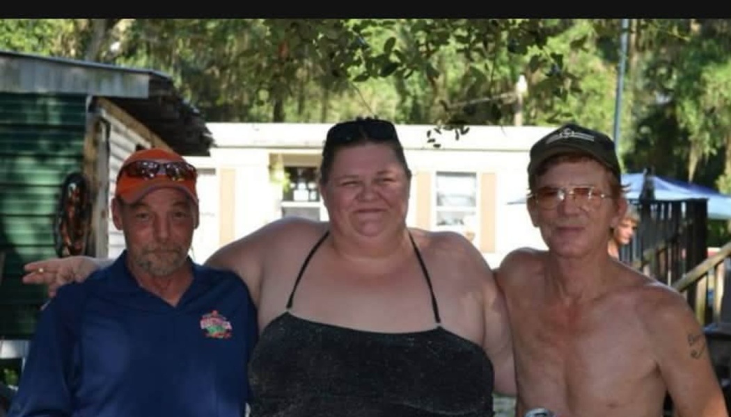 A smiling woman with her arms around two men outdoors in front of a trailer home.