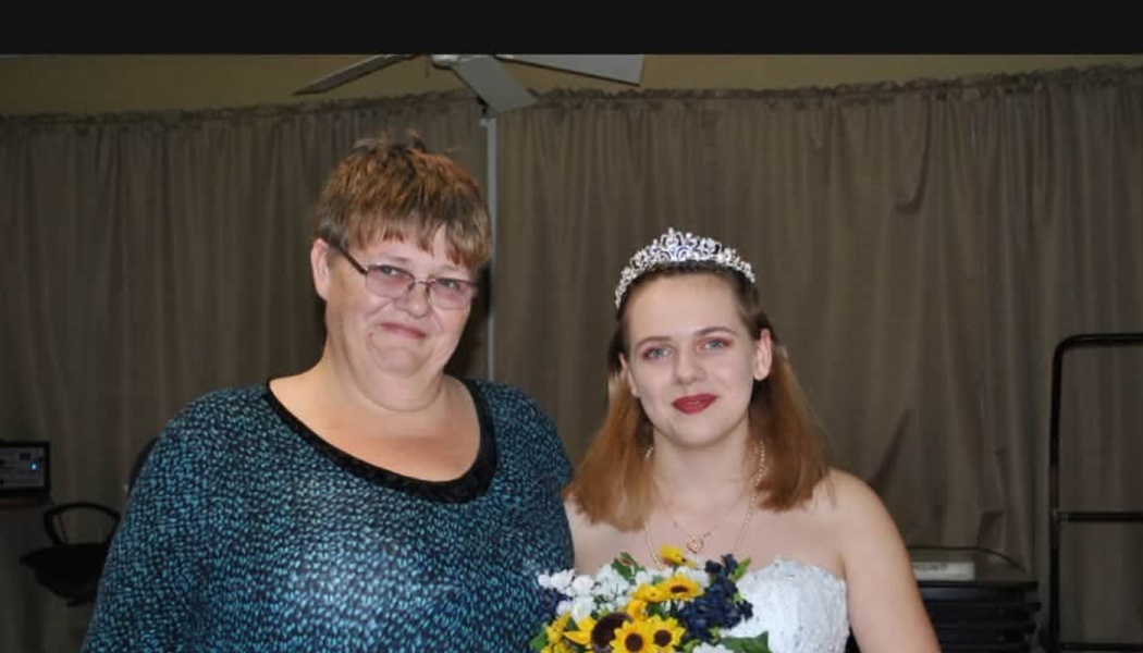 A young woman wearing a tiara and white dress holding a bouquet stands smiling beside an older woman in glasses.
