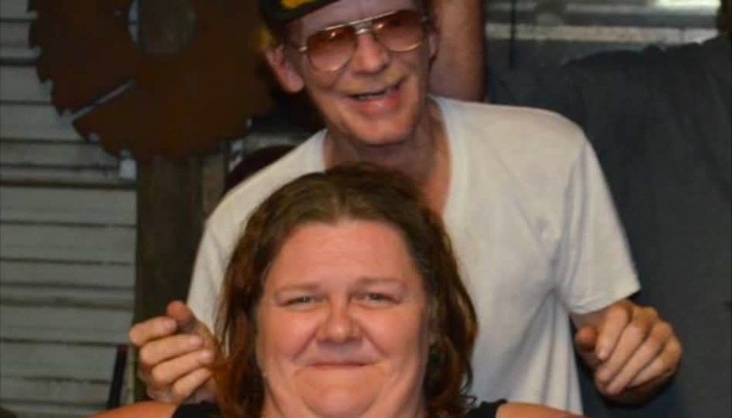A smiling man wearing tinted glasses stands behind a smiling woman with brown hair.