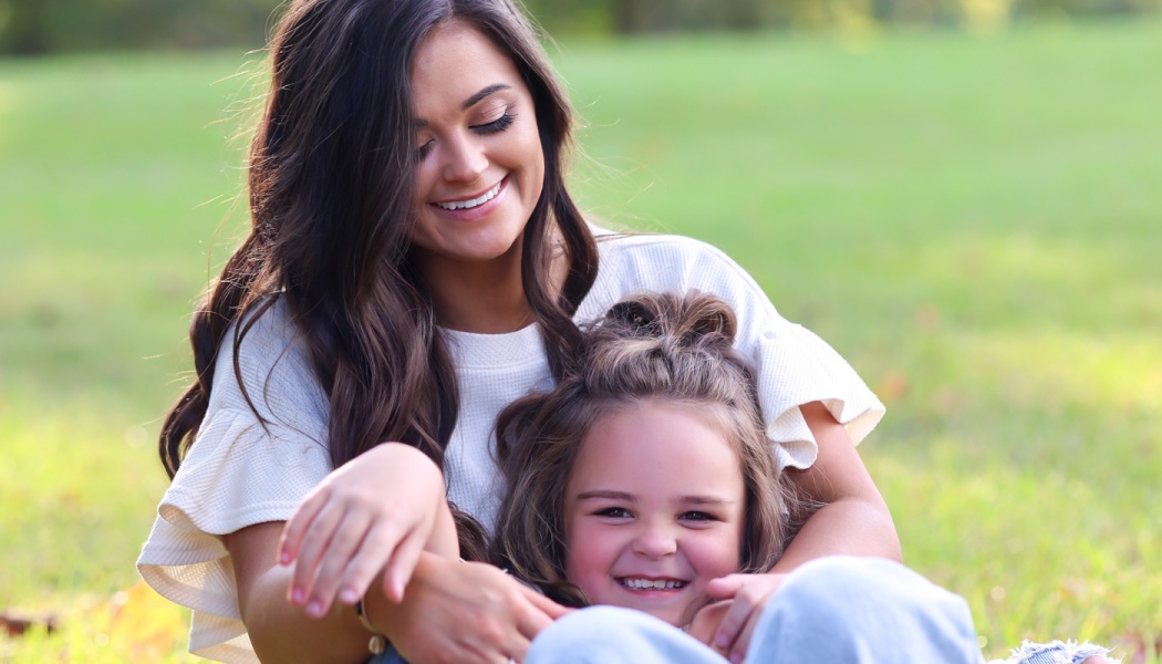 A smiling woman with long dark hair lovingly embraces a happy young girl outdoors on a grassy lawn.