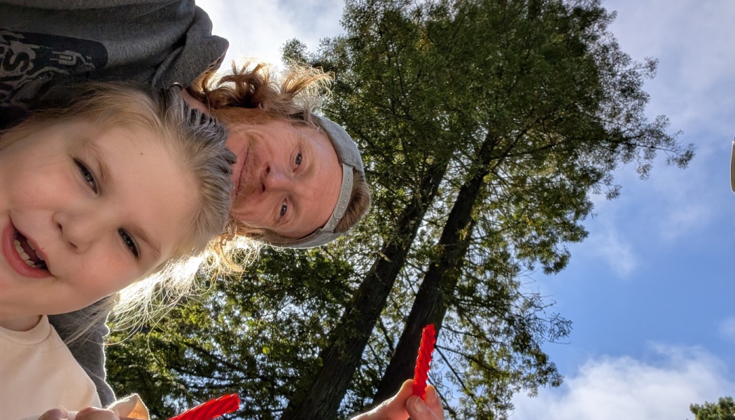 A smiling man and young girl holding red licorice outdoors, framed by tall trees and blue sky.
