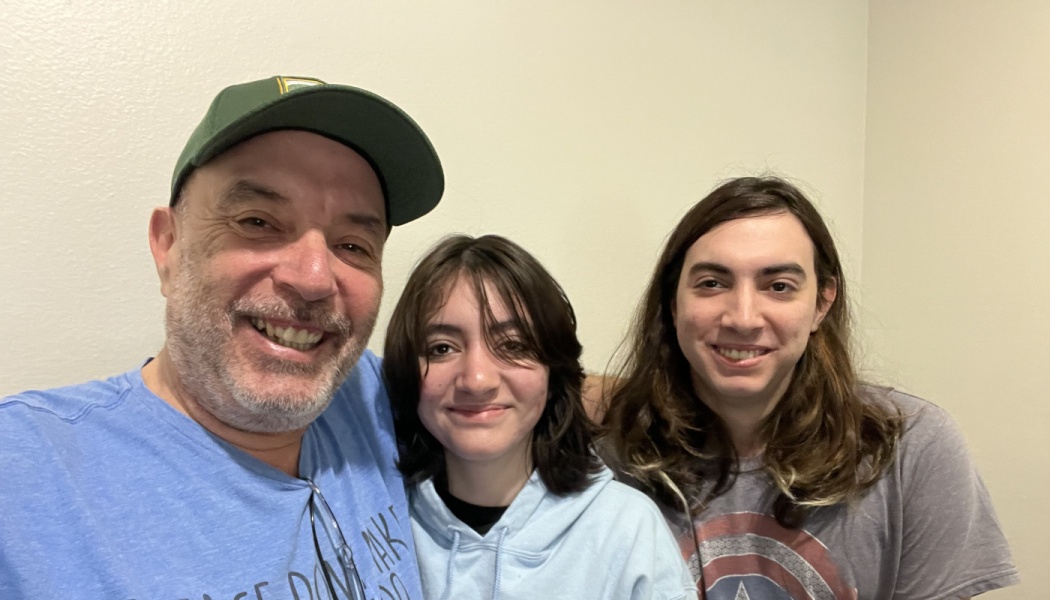 A smiling man in a green cap poses for a close selfie with two young adults against a neutral wall.