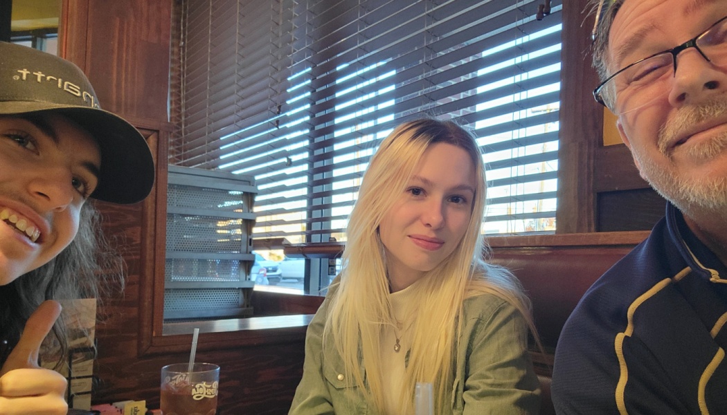 Three people smiling for a selfie while sitting in a restaurant booth next to window blinds.