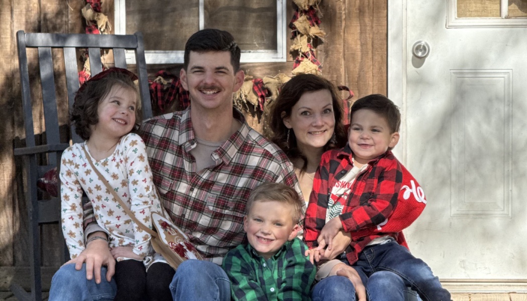 A smiling family of five with three young children posing together on the porch of a rustic wooden cabin.