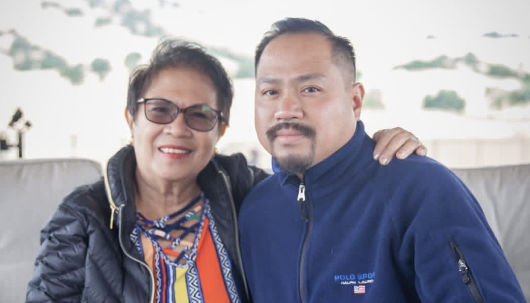 A smiling woman with glasses rests her hand on the shoulder of a man with a goatee wearing a blue jacket.