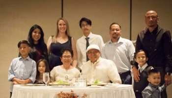 A multi-generational family smiling together around an elderly couple seated at a formal dinner table.