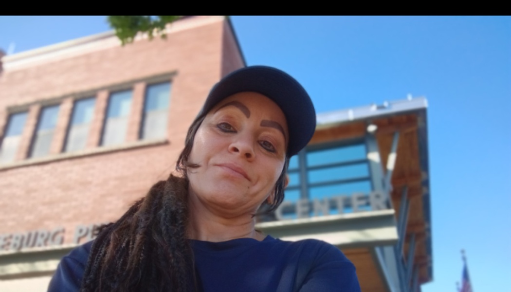 A woman wearing a dark cap takes a low-angle selfie outdoors in front of a brick building on a clear, sunny day.