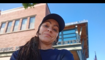 A woman wearing a dark cap takes a low-angle selfie outdoors in front of a brick building on a clear, sunny day.