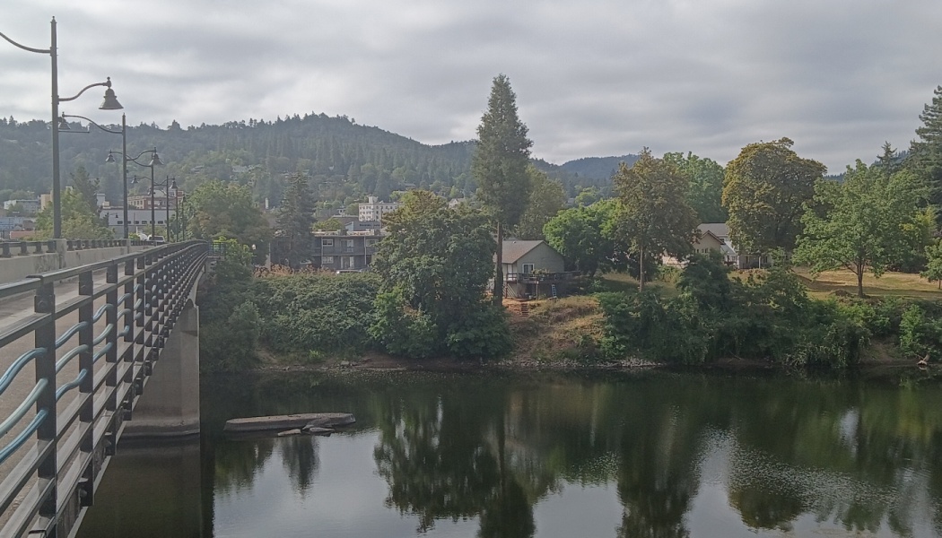 Bridge railing overlooking a calm river reflecting lush trees and houses, with forested hills in the background.
