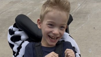 A young blond boy with a wide, joyful smile sitting in a wheelchair.