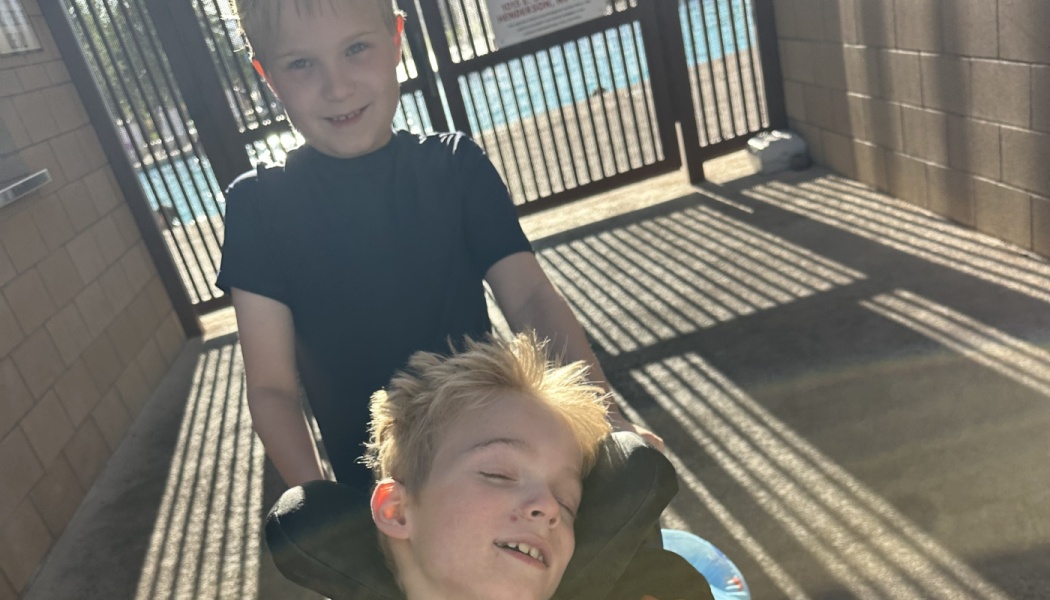 A smiling boy stands behind a blonde boy resting comfortably in a wheelchair outdoors.