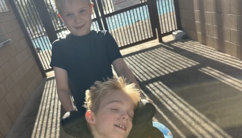 A smiling boy stands behind a blonde boy resting comfortably in a wheelchair outdoors.