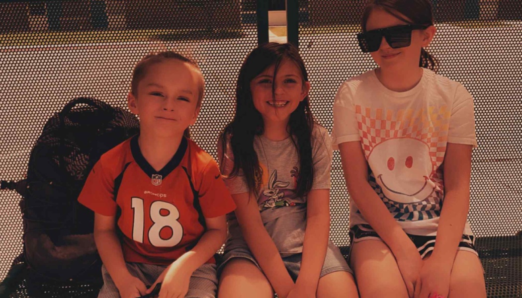 Three happy young children sitting side-by-side on an outdoor bench, smiling at the camera.