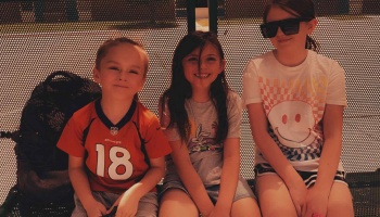 Three happy young children sitting side-by-side on an outdoor bench, smiling at the camera.