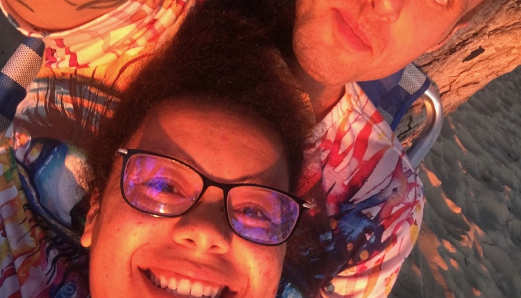 A smiling woman with glasses resting beside a man outdoors under warm golden sunset lighting.