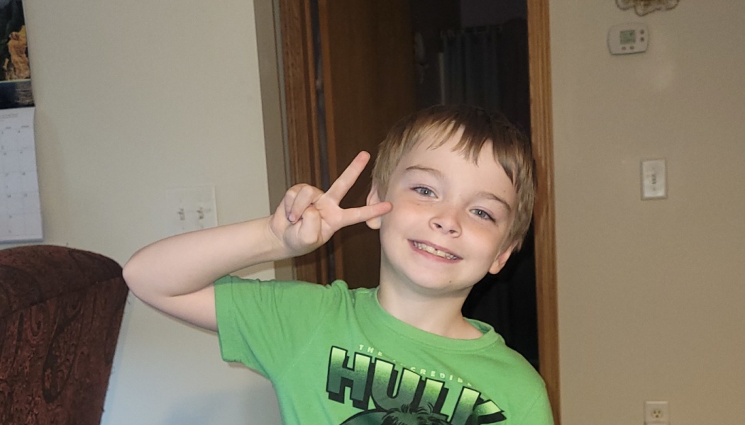 A smiling young boy wearing a green Hulk shirt giving a peace sign.