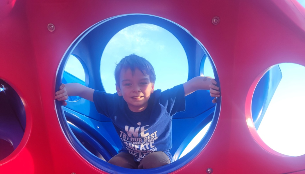 A smiling young boy in a blue shirt looking through a circular opening in a red and blue playground structure.