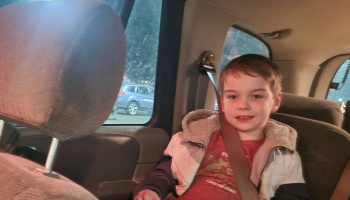 A young boy wearing a jacket sits buckled into the back seat of a car, smiling at the camera.