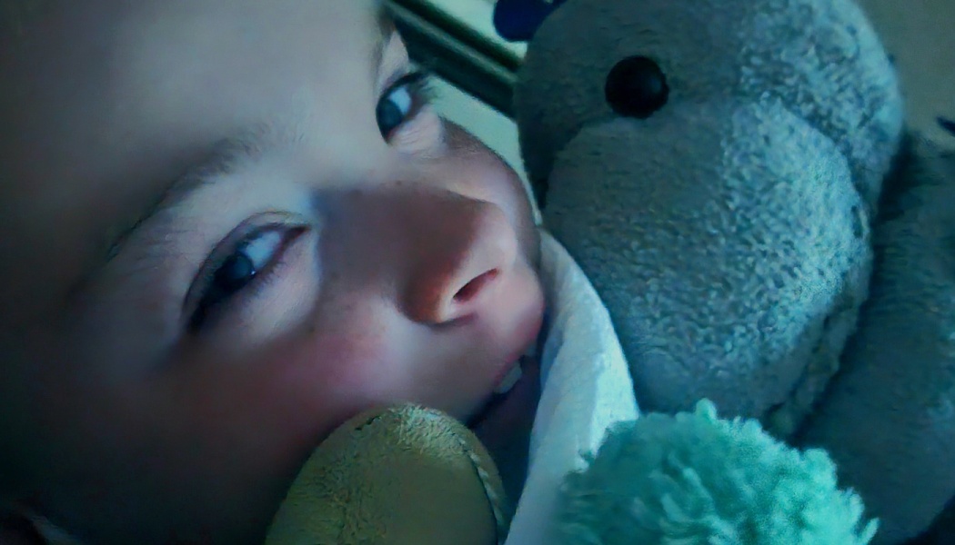Close-up of a smiling child snuggled in bed next to a plush stuffed animal.