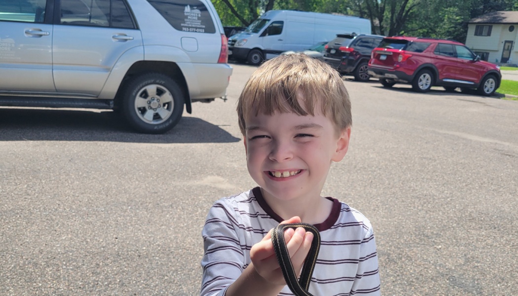 A smiling young boy in a striped shirt proudly holds a small garter snake outdoors.