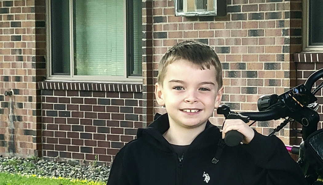 A smiling young boy in a black hoodie holding the handlebars of a bicycle outside a brick building.