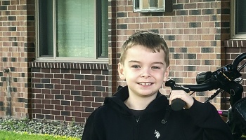 A smiling young boy in a black hoodie holding the handlebars of a bicycle outside a brick building.