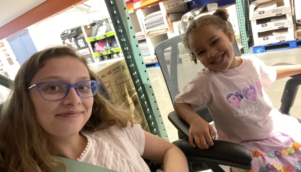 A woman with blue glasses takes a selfie with a smiling young girl sitting in an office chair inside a store.