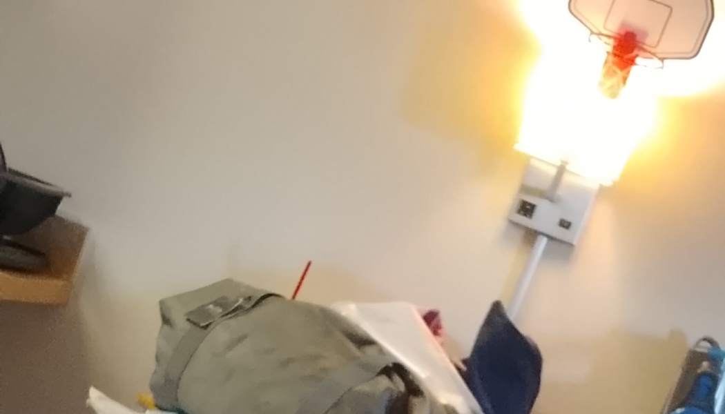 Green duffel bag sitting in a room near a bright wall lamp with a toy basketball hoop attached above it.
