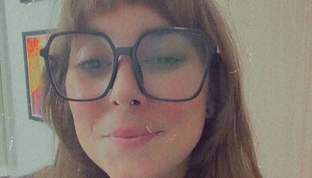 Close-up portrait of a smiling woman with bangs wearing large, black-framed glasses.