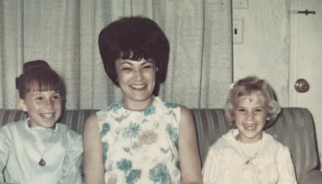 Smiling mother with a 1960s beehive hairstyle sitting on a couch between her two happy young daughters.