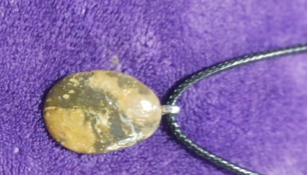 Smooth, marbled brown and tan oval stone pendant attached to a black cord necklace on a plush purple fabric.