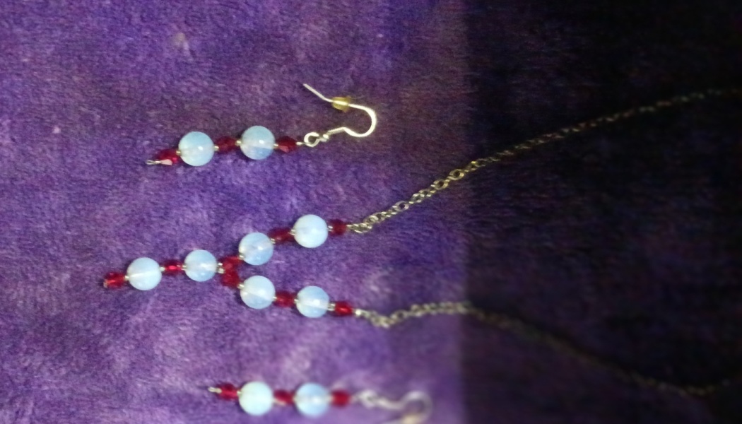 Handcrafted jewelry set featuring light blue opalescent beads and red crystals on a purple velvet backdrop.