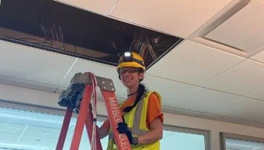A smiling female electrician in a hard hat with a headlamp stands on a ladder working on ceiling wiring.