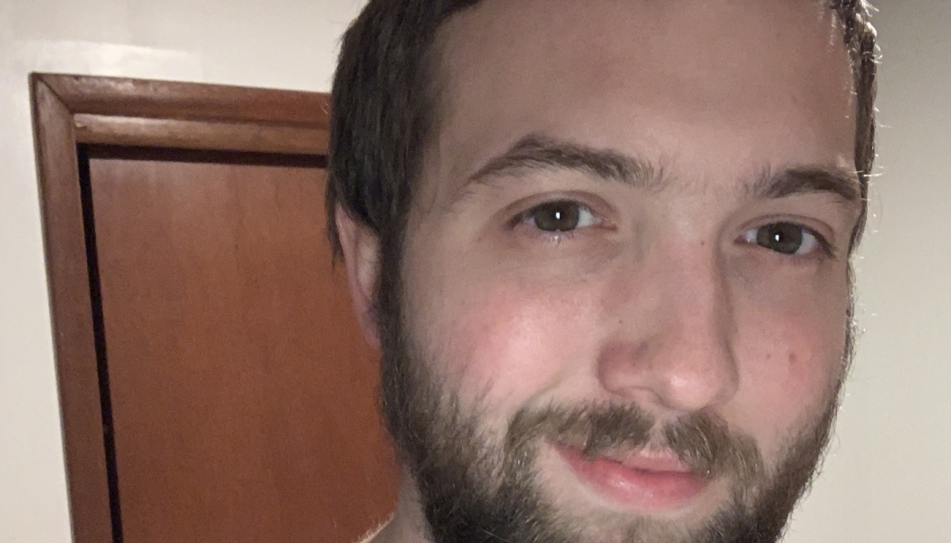 Close-up selfie of a young man with short brown hair, a beard, and brown eyes, smiling slightly.