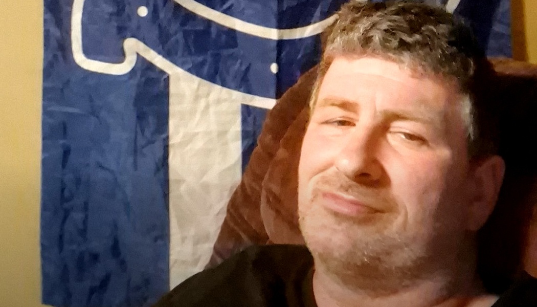 A man with short graying hair and stubble smiling slightly, sitting in front of a blue and white flag.