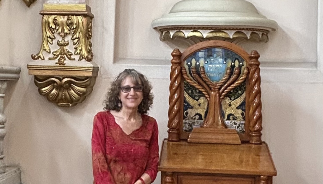 A smiling woman in a red shirt stands next to an ornate wooden lectern featuring a menorah mosaic.